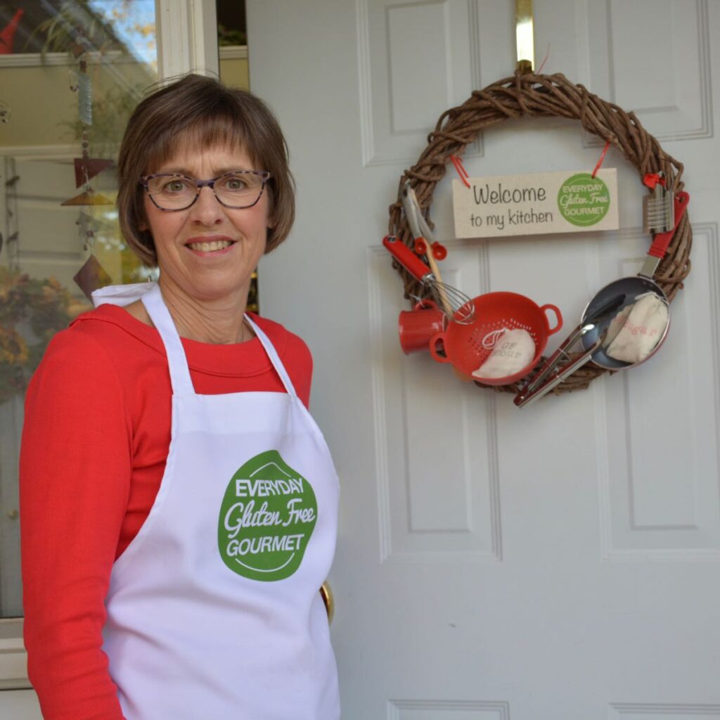 Cinde wearing her EGFG apron, standing outside her front door with a kitchen gadget wreath that includes a sign saying 'Welcome to my kitchen.'