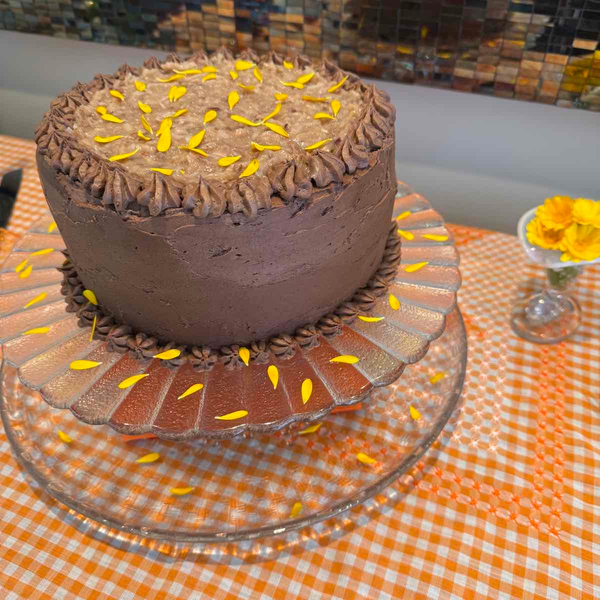 A three layer German Chocolate Cake with coconut-pecan filling on a pedestal tray. Covered with chocolate icing and edible calendula petals.