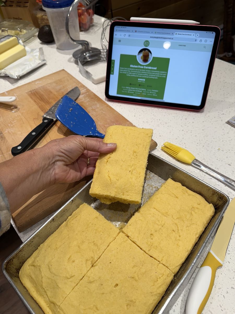A kitchen counter with recipe on the iPad and a pan of cornbread cut in four, with someone holding one piece.