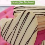 A stack of heart shaped gluten free sugar cookies drizzled with chocolate sitting on pretty pink paper surrounded by tiny heart shaped sprinkles.