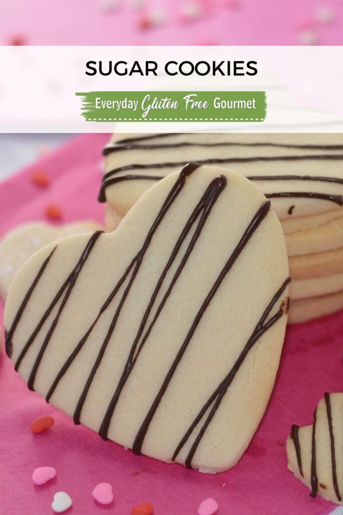 A stack of heart shaped gluten free sugar cookies drizzled with chocolate sitting on pretty pink paper surrounded by tiny heart shaped sprinkles.