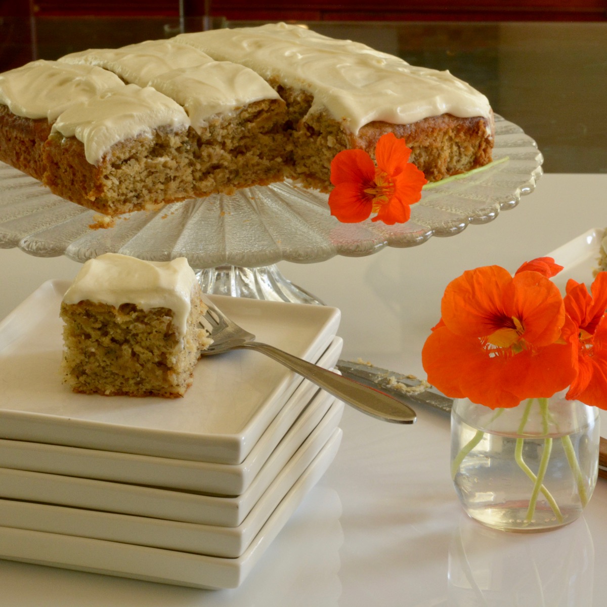 A banana cake with cream cheese icing on a pedestal tray with a single piece on a plate ready to serve.