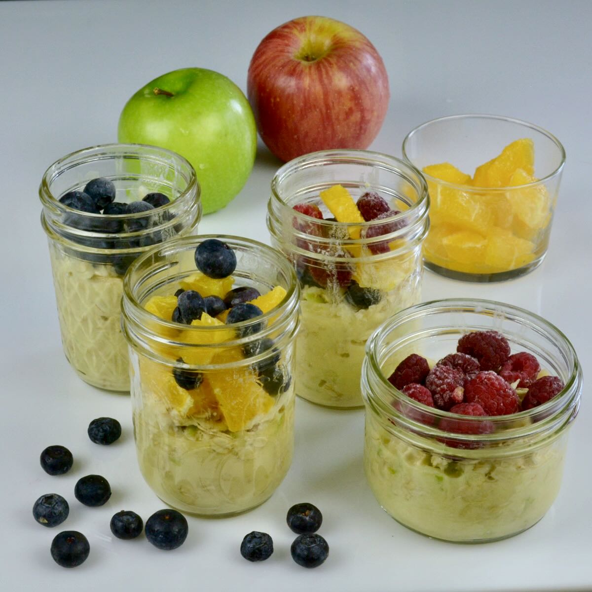 Four jars of overnight oats topped with oranges, blueberries and raspberries surrounded by whole apples and a few blueberries.